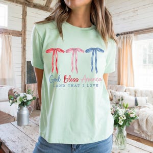 May include: A light green t-shirt with three red, white and blue bows and the text "God Bless America Land That I Love".