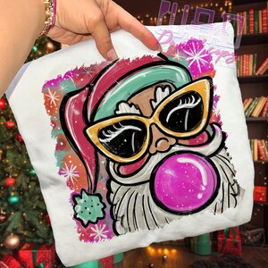 May include: White Christmas-themed sweatshirt featuring a cartoon Santa Claus with yellow sunglasses and a pink bubble. The Santa Claus has a red and green hat and a white beard. The design is surrounded by colorful snowflakes.
