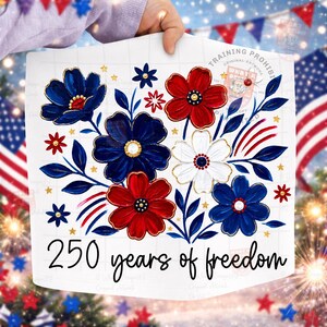 May include: A white print featuring red, white, and blue floral designs with gold accents. The text "250 years of freedom" is written in a script font. The design is held up by a person.