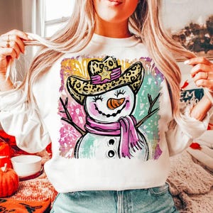 May include: White sweatshirt featuring a whimsical snowman design. The snowman wears a leopard print cowboy hat, a pink scarf, and has a carrot nose. The background has colorful brushstrokes of pink, yellow, and teal.