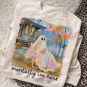 K&ouml;nnte beinhalten: Ein cremefarbenes Sweatshirt mit Rundhalsausschnitt und einem Grafikdruck eines Geistes, der auf einem Stuhl vor einem Haus mit orangefarbenen Herbstb&auml;umen sitzt. Der Geist h&auml;lt einen Kaffeebecher und der Text lautet "mentally i'm here."