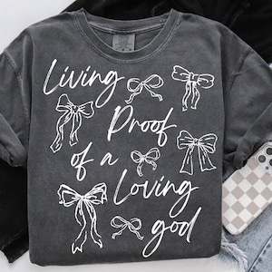 May include: A dark grey t-shirt with white text that reads "Living Proof of a Loving Good" and several white bow designs.