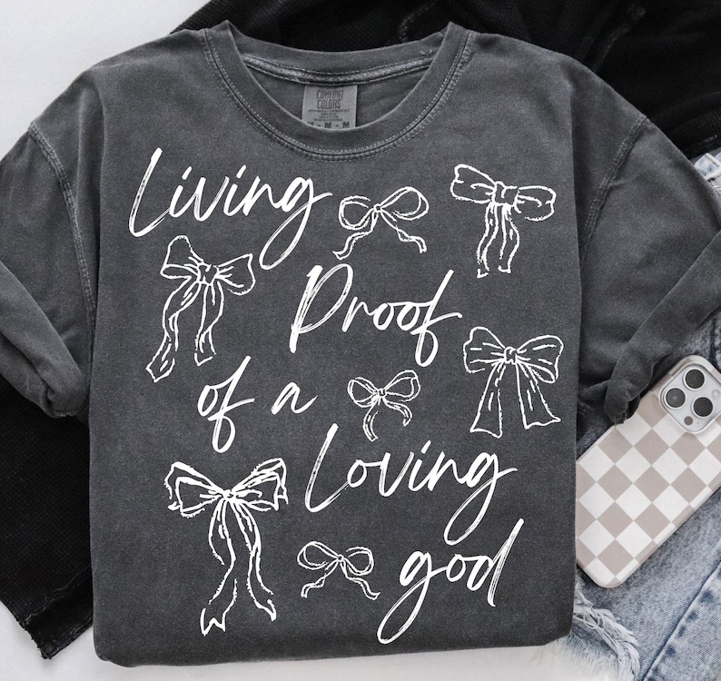 May include: A dark grey t-shirt with white text that reads "Living Proof of a Loving Good" and several white bow designs.