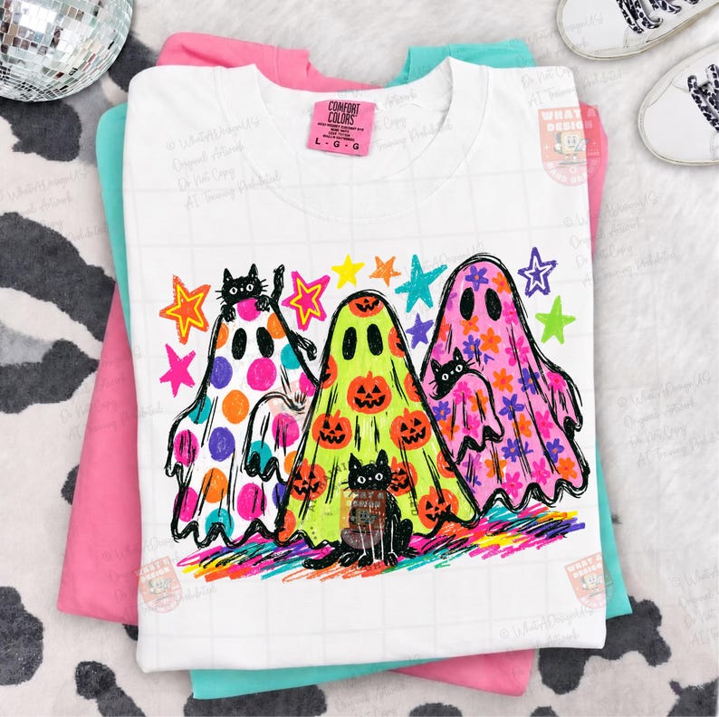 K&ouml;nnte beinhalten: Ein wei&szlig;es T-Shirt mit einer Grafik von drei Geistern in bunten Mustern. Ein Geist hat orangefarbene Punkte, einer hat orangefarbene K&uuml;rbisse auf hellgr&uuml;nem Hintergrund und einer hat rosa Blumenmuster. Schwarze Katzen sind im Design integriert.