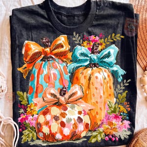 May include: A black t-shirt featuring a graphic of three pumpkins decorated with large bows. The pumpkins are orange and blue with polka dot patterns, surrounded by colorful floral accents in shades of pink, yellow, and green.