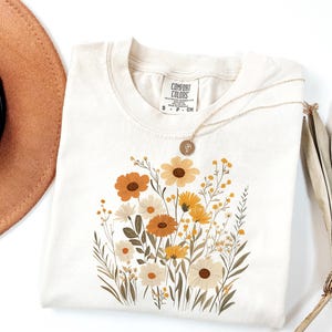 May include: A beige t-shirt with a floral print featuring yellow, orange, and brown flowers. The shirt has a tag that reads "Comfort Colors" and "S - P - CH".