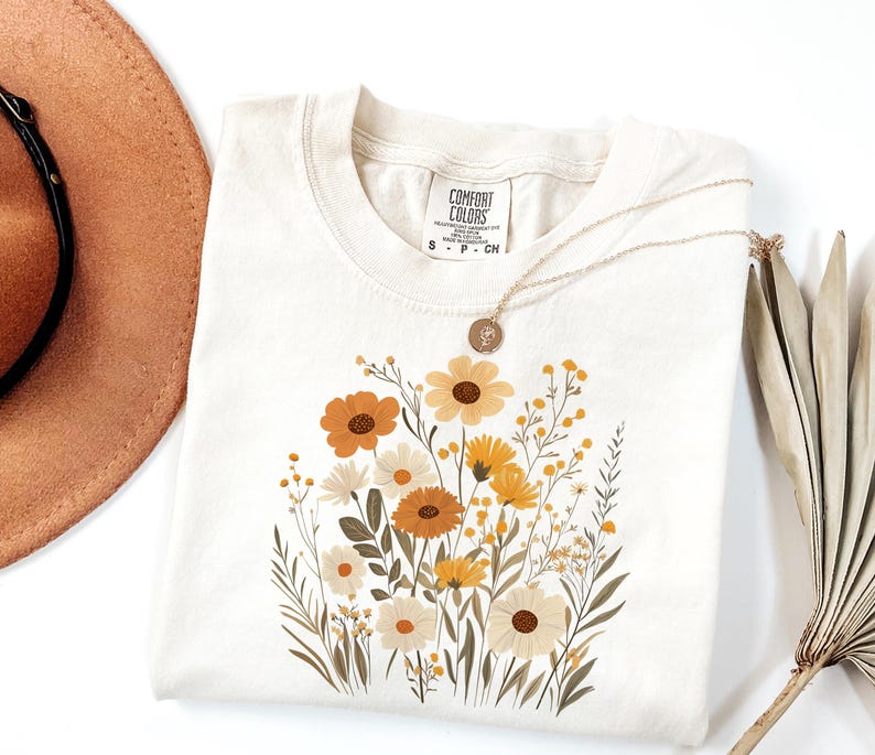 May include: A beige t-shirt with a floral print featuring yellow, orange, and brown flowers. The shirt has a tag that reads "Comfort Colors" and "S - P - CH".