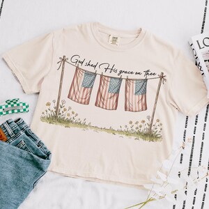 May include: Cream-colored t-shirt with the text "God shed His grace on thee" above a graphic of three American flags hanging on a clothesline. The shirt is paired with blue jeans and a green and white checkered hair clip.