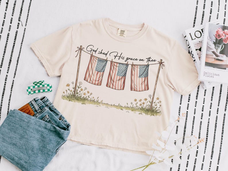 May include: Cream-colored t-shirt with the text "God shed His grace on thee" above a graphic of three American flags hanging on a clothesline. The shirt is paired with blue jeans and a green and white checkered hair clip.