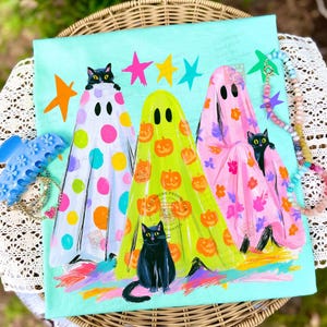 May include: A light blue t-shirt featuring a graphic of three ghosts in white, yellow, and pink, each decorated with colorful polka dots, pumpkins, and floral patterns. Three black cats are positioned among the ghosts, with colorful stars above.