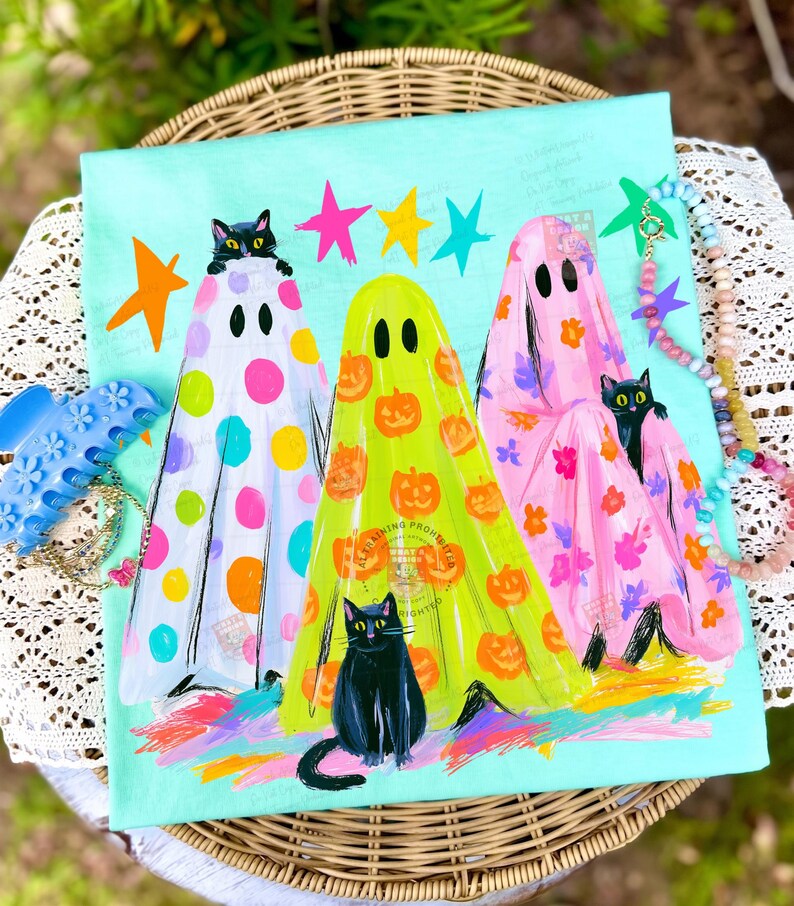 May include: A light blue t-shirt featuring a graphic of three ghosts in white, yellow, and pink, each decorated with colorful polka dots, pumpkins, and floral patterns. Three black cats are positioned among the ghosts, with colorful stars above.