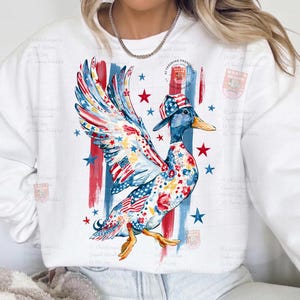 May include: A white sweatshirt with a colourful duck illustration. The duck is decorated with patriotic patterns, including the American flag, stars, and stripes. The design incorporates red, white, and blue colours, with the text "AT TRAINING PROHIBITED".