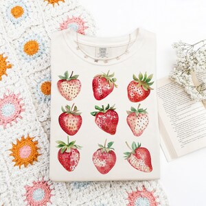 Op de afbeelding: Een wit T-shirt met een aquarelontwerp van aardbeien. De aardbeien zijn rood met groene bladeren en zijn gerangschikt in een rasterpatroon.