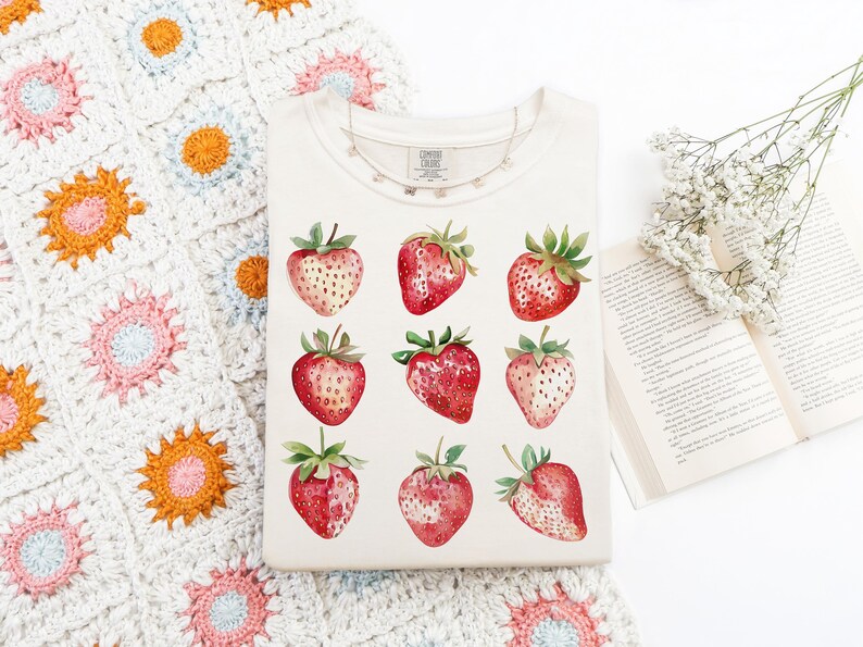 Op de afbeelding: Een wit T-shirt met een aquarelontwerp van aardbeien. De aardbeien zijn rood met groene bladeren en zijn gerangschikt in een rasterpatroon.
