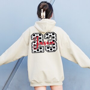 May include: A beige hoodie with a black and white soccer ball design on the back. The design includes the number 23 and the name "Jassen" in red.