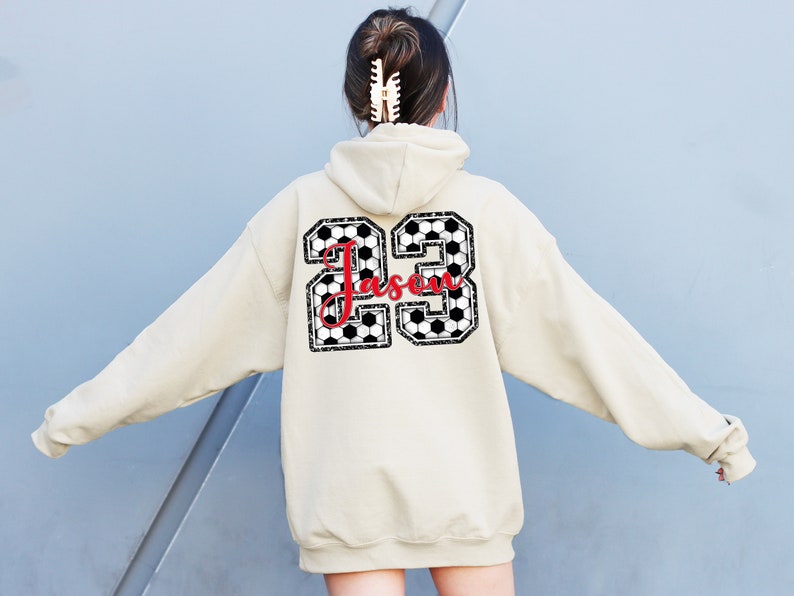May include: A beige hoodie with a black and white soccer ball design on the back. The design includes the number 23 and the name "Jassen" in red.