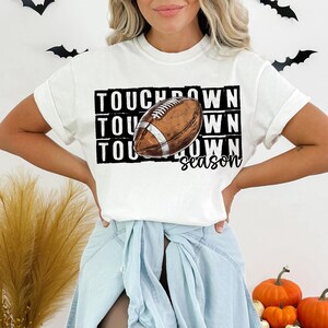 Op de afbeelding: Wit T-shirt met een zwart-witte afbeelding van een voetbal en de tekst "Touchdown season".