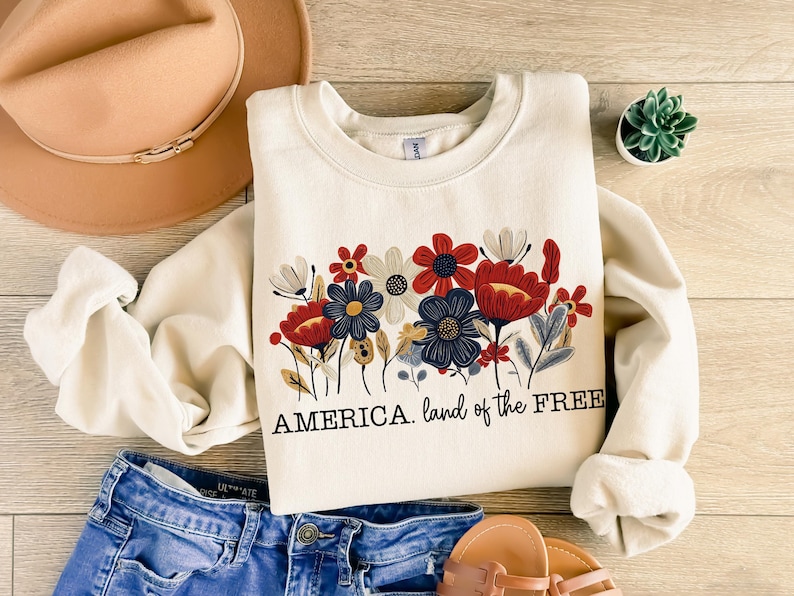 May include: A white sweatshirt with a floral design in red, blue, and white. The text "AMERICA. land of the FREE" is printed below the flowers.