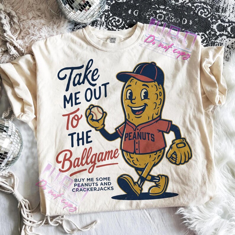 May include: Cream-colored t-shirt with the text "Take Me Out To The Ballgame" and a cartoon peanut character in a baseball uniform. The peanut is holding a baseball and wearing a cap. The shirt also says "Buy me some peanuts and crackerjacks."
