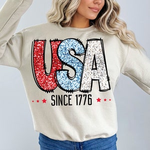 May include: A beige sweatshirt with a red, white, and blue glitter graphic that says "USA SINCE 1776" with red stars on either side of the text.
