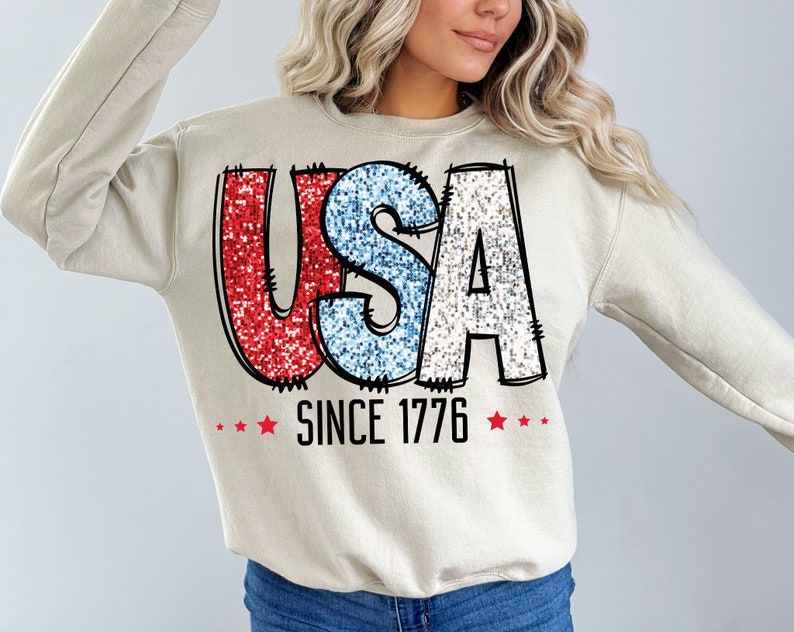 May include: A beige sweatshirt with a red, white, and blue glitter graphic that says "USA SINCE 1776" with red stars on either side of the text.