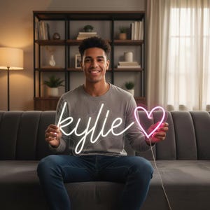 Könnte beinhalten: Ein Neon-Schild mit dem Namen "Kylie" und einem Herz. Der Name ist in weißer Schreibschrift, das Herz ist rosa. Die Person hält das Schild. Im Hintergrund sind ein Sofa und ein Bücherregal zu sehen.