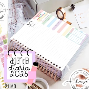 Puede incluir: Una agenda diaria con espiral y esquema de colores pastel. La agenda tiene un interior punteado, pestañas etiquetadas y un calendario de mayo. La portada dice "agenda diaria 2026". Accesorios: pegatinas, bolígrafo dorado y clips.