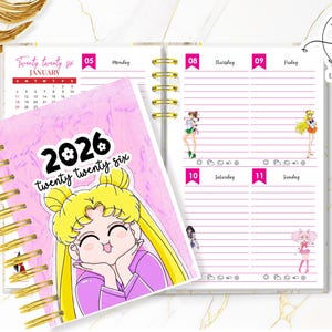 Puede incluir: Una agenda rosa de 2026 con un personaje de dibujos animados en la portada. La agenda tiene encuadernación en espiral dorada y presenta diseños semanales con ilustraciones. La portada dice "twenty twenty six".
