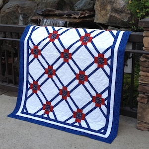 May include: A white quilt with a blue and red geometric pattern. The quilt is folded and draped over a railing in front of a waterfall.