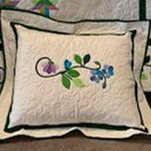 May include: A white pillow with a green vine and purple flowers embroidered on it. The pillow has a white border and a green trim.