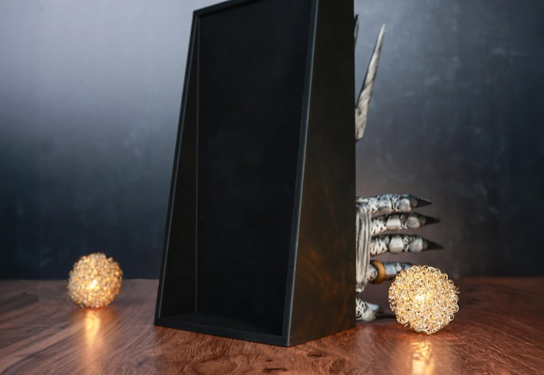 Puede incluir: Una caja de exhibici&oacute;n de madera negra con un frente en &aacute;ngulo, colocada sobre una superficie de madera. Dos luces decorativas esf&eacute;ricas de color dorado se colocan cerca de la caja. Tambi&eacute;n se ve un fondo oscuro y una mano decorativa con garras.