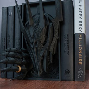 Puede incluir: Un sujetalibros negro con un dise&ntilde;o de drag&oacute;n estilizado y una mano que sostiene un anillo dorado. Se encuentra entre libros con t&iacute;tulos como "HAPPY SEXY MILLIONAIRE" y "THINK AND GROW RICH".