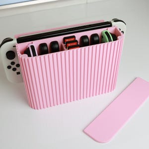 May include: Pink Nintendo Switch console storage case with a white console and two controllers inside. The case has a ribbed texture and a separate lid.