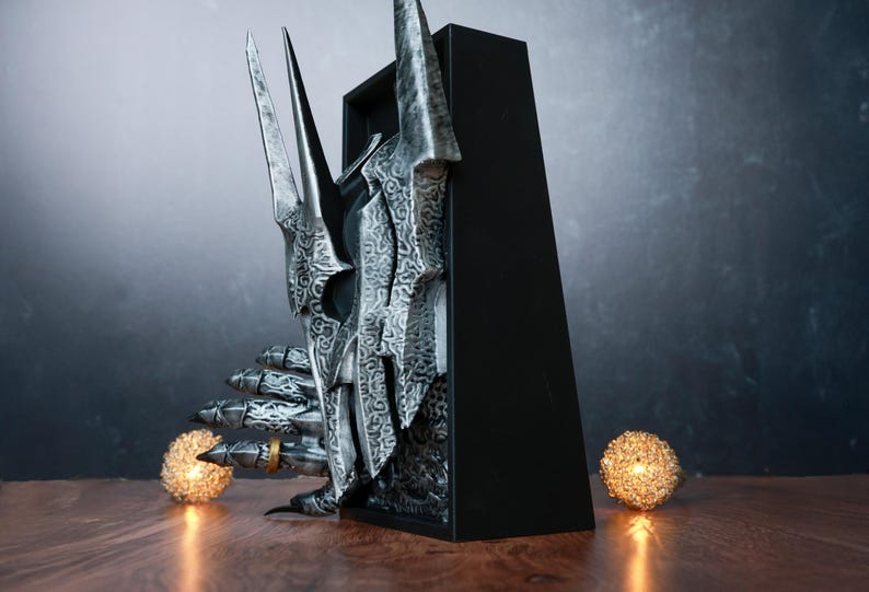 Puede incluir: Una detallada escultura plateada de un casco y una mano de personaje de fantas&iacute;a, con intrincados patrones y bordes afilados. La escultura se encuentra sobre un fondo oscuro, con un anillo dorado en un dedo. Dos orbes brillantes flanquean la escultura.