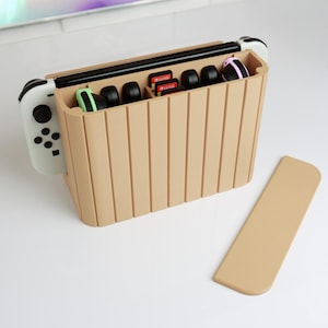 Könnte beinhalten: Ein beigefarbener Aufbewahrungskasten aus Kunststoff für eine Nintendo Switch-Konsole, zwei Controller und Spielpatronen. Der Kasten hat einen Einschub für die Konsole, zwei Einschübe für Controller und ein Fach für Spielpatronen. Der Kasten ist geöffnet und die Konsole, Controller und Patronen sind sichtbar.