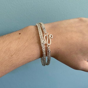 Könnte beinhalten: Zwei silberne Armbänder mit einem geflochtenen Kettendesign. Die Armbänder werden am Handgelenk getragen und haben einen silbernen Verschluss.
