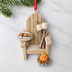 May include: A tan wooden Adirondack chair ornament featuring a plate with three s'mores, two marshmallows on sticks, and a small campfire at the base. The ornament hangs from a red ribbon and includes a green pine branch at the top.