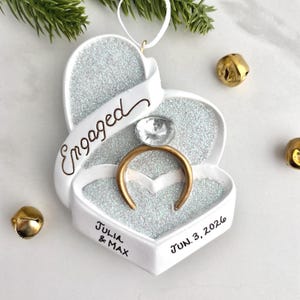 May include: A heart-shaped white ornament featuring a gold ring with a clear stone, silver glitter accents, and the text "Engaged", "Julia & Max", and "JUN. 3, 2026". The ornament includes a white hanging string and is surrounded by gold jingle bells.