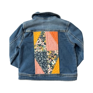 May include: A denim jacket with a patchwork design on the back. The patchwork features floral and geometric patterns in shades of blue, yellow, orange, and pink. The jacket has a classic collar and button closures.