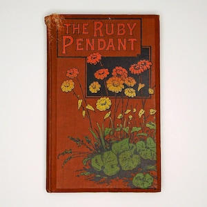 Peut inclure: Un livre vintage intitulé "The Ruby Pendant" avec une couverture brun-rouge. La couverture présente une illustration florale avec des accents orange, jaune et vert. Le titre est en lettres orange en gras. Les bords du livre montrent des signes d'usure.
