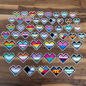May include: A collection of 40 pixelated heart stickers featuring various pride flags, including the rainbow, transgender, bisexual, pansexual, and non-binary flags. The stickers are arranged in rows on a wooden surface.