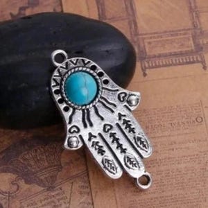 Può includere: Ciondolo in argento a forma di mano di hamsa con una pietra turchese al centro. La mano di hamsa è un simbolo di protezione e buona fortuna.