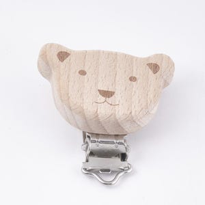 Peut inclure: Un clip en bois en forme d'ours avec un fermoir métallique. L'ours a un visage simple dessiné à la main avec deux yeux, un nez et une bouche.