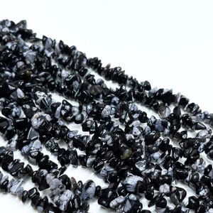 May include: Close-up of several strands of black and white snowflake obsidian chip beads. The beads are irregularly shaped and have a polished, glossy finish. The background is plain white.