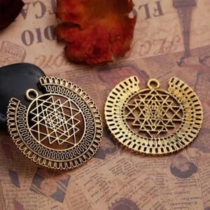 May include: Two antique gold-colored pendants with intricate geometric designs. Each pendant features a central pattern of intersecting lines and shapes, surrounded by a decorative border. The pendants have a loop at the top for hanging.