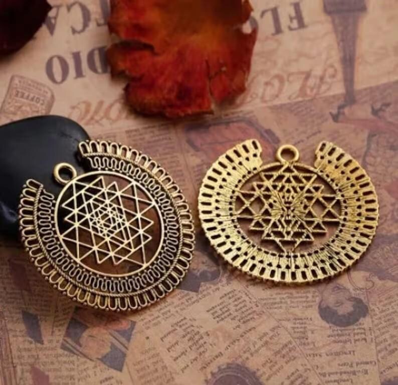 May include: Two antique gold-colored pendants with intricate geometric designs. Each pendant features a central pattern of intersecting lines and shapes, surrounded by a decorative border. The pendants have a loop at the top for hanging.