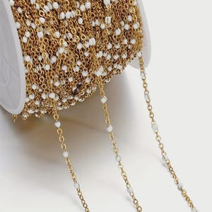 Peut inclure: Une chaîne dorée avec des perles blanches, idéale pour la fabrication de bijoux. La chaîne est enroulée sur une bobine blanche et une partie de la chaîne est disposée en ligne droite.