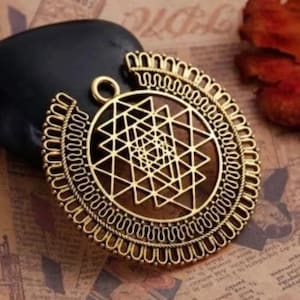 May include: A gold-colored pendant with a geometric design. The pendant features a central geometric pattern within a circular frame, with intricate detailing. The pendant is likely made of metal and has a loop for hanging.