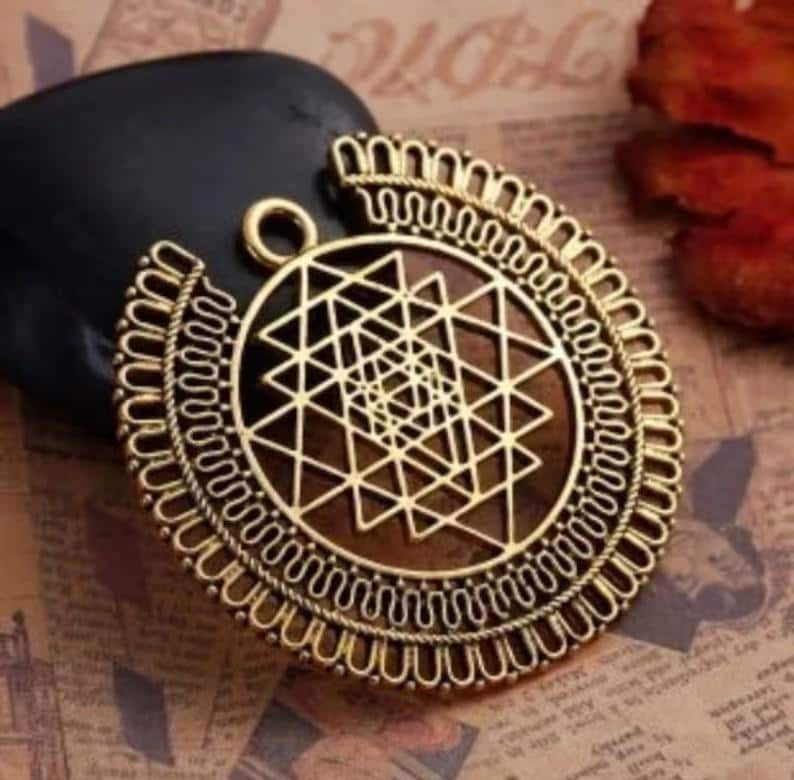 May include: A gold-colored pendant with a geometric design. The pendant features a central geometric pattern within a circular frame, with intricate detailing. The pendant is likely made of metal and has a loop for hanging.