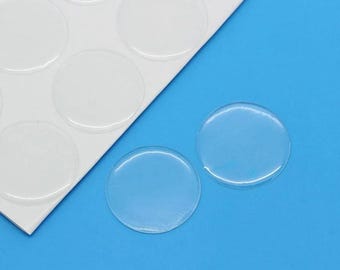 Può includere: Un foglio di adesivi rotondi adesivi trasparenti su un supporto bianco. Due adesivi singoli sono mostrati su una superficie blu.